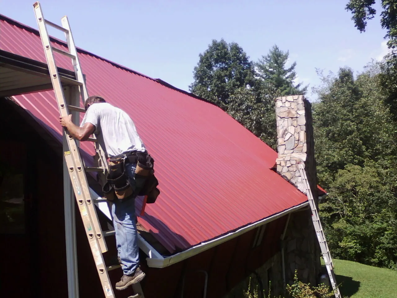 Roofer on a ladder beside a red metal roof and stone chimney — Soffit & Fascia Repair by Roofing Brothers in New Palestine